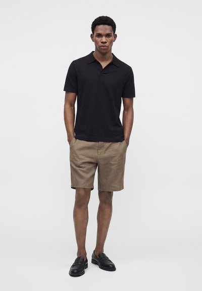 Homme debout les mains dans les poches, portant un polo noir à manches courtes, un short beige et des mocassins noirs sur un fond uni.