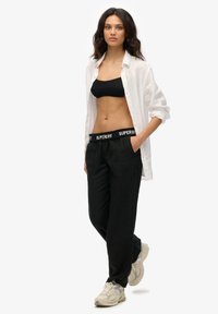 Pantalons en lin noirs avec une ceinture élastique marquée, associés à un bralette en tricot noir et une chemise blanche ouverte. Des baskets beiges complètent le look.