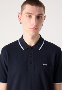 Navy polo majica iz bombaža, z zapenjanjem na tri gumbke, kontrastnim svetlo modrim ovratnikom in obrobami rokavov ter majhnim belim logotipom.
