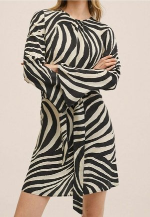 Femme portant une robe à manches longues rayée zèbre noire et blanche avec une ceinture à la taille, debout, les bras croisés.