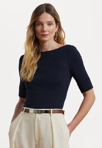 Chemise ajustée bleu marine à manches trois-quarts et décolleté bateau, associée à un pantalon beige taille haute et une ceinture avec des accents argentés.