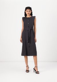 Vestido sem mangas de tom cinza escuro com corpete ajustado, botões na frente, faixa na cintura e saia em forma de A. Combinado com sandálias pretas de salto alto com detalhes.