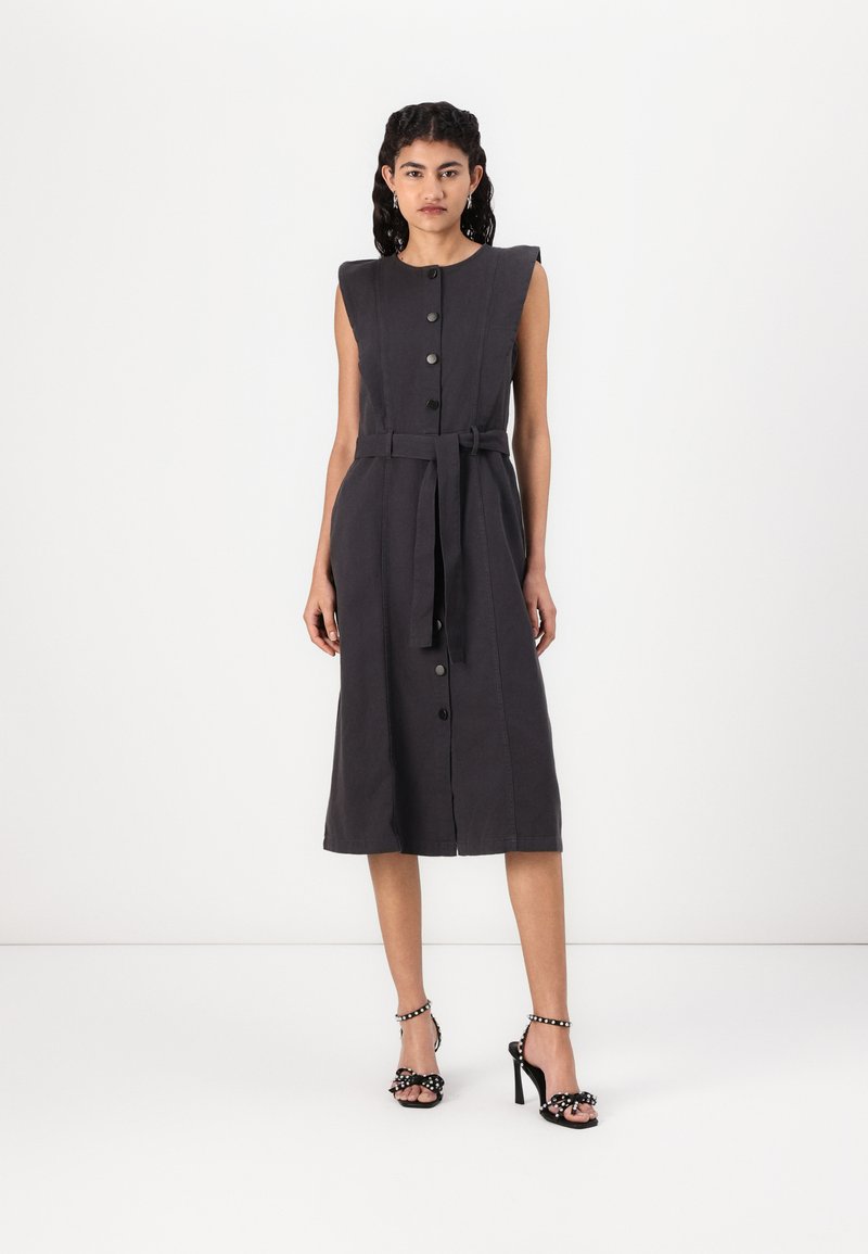 Vestido sem mangas de tom cinza escuro com corpete ajustado, botões na frente, faixa na cintura e saia em forma de A. Combinado com sandálias pretas de salto alto com detalhes.