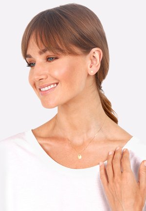 Gold necklace with a circular pendant, paired with gold stud earrings. The model wears a white shirt, showcasing a light skin tone.