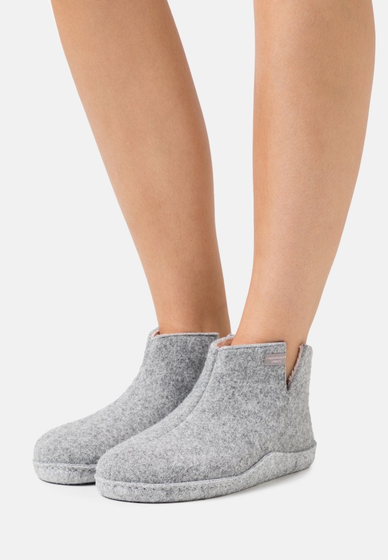 Pieds portant des chaussons chevilles en feutre gris clair avec une doublure douce et une petite étiquette en tissu sur le côté extérieur, sur un fond uni.