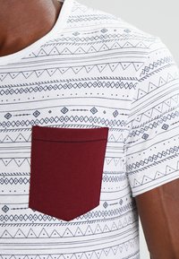 White T-shirt with geometric blue patterns, round neckline, and a burgundy pocket on the left. Fabric appears soft with a smooth texture.