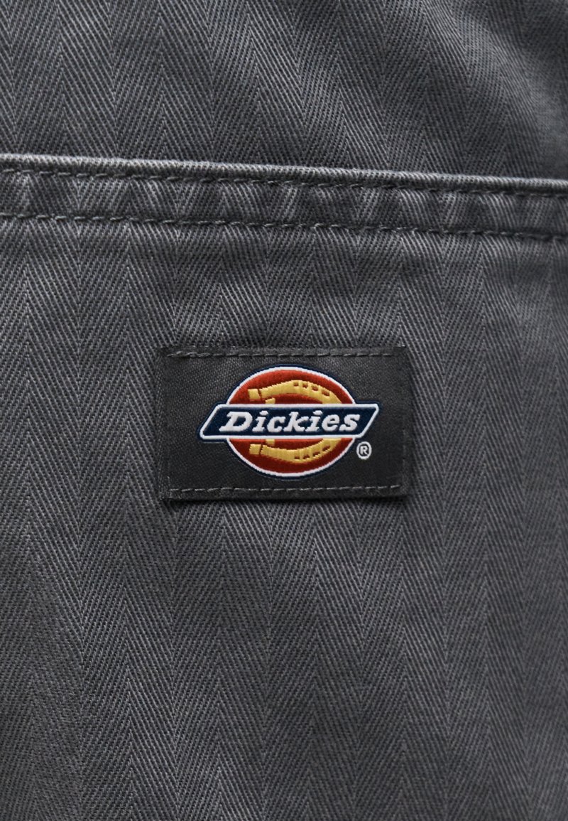 Dickies merklabel genaaid op grijze stof met een visgraatpatroon en zichtbare dubbele stiksel boven het label.