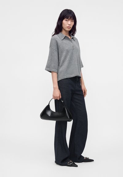Femme portant un polo gris à manches courtes, pantalon large foncé, chaussures noires pointues, tenant un sac à main noir sur un fond uni.