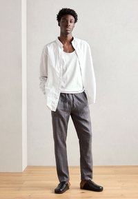 White striped button-up shirt over a white tank top, paired with gray drawstring trousers and black slip-on shoes. Wood floor background.