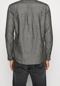 Uomo che indossa una camicia grigia con maniche lunghe e jeans grigio scuro, visto da dietro su uno sfondo neutro.