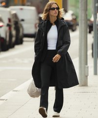 Cămașă lungă neagră peste un top alb, colanți negri și adidași albi. Ochelari de soare negri. Mediu urban exterior cu un trotuar.