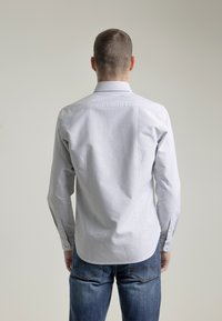 Camisa de manga larga, con botones, en azul claro con finas rayas blancas, que presenta un cuello clásico y botones en los puños. Llevada con jeans azules.