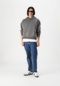 Hanorac gri cu glugă și buzunar frontal, purtat peste o cămașă albă. Blugi din denim albastru și pantofi sport cu accente negre.