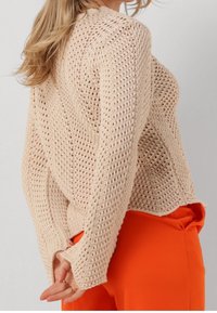 Pull beige en maille avec un motif à tissage ouvert et manches longues, porté sur un pantalon orange vif. Il présente un col rond et une coupe ample.