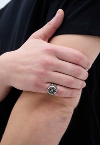 Anel de prata com um design de estrela preta, usado numa mão que envolve um braço. O anel apresenta detalhes intrincados e texturas contrastantes.