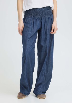 Osoba nosiaca voľné modré rifľové nohavice so širokými nohavicami a elastickým nariaseným pásom a béžové ležérne topánky, stojaca na jednoduchom pozadí.