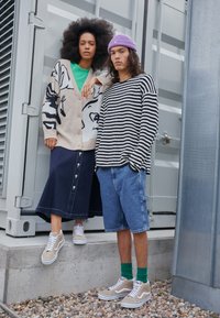 Cardigan beige avec imprimé floral noir, t-shirt vert, longue jupe en jean à boutons ; pull rayé noir et blanc, shorts cargo, baskets beiges.