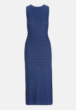 Žena u plavoj pamučnoj midi haljini bez rukava, zlatnoj ogrlici, zlatnoj narukvici i satu, drži zlatnu torbicu, stoji nasuprot bijeloj pozadini.