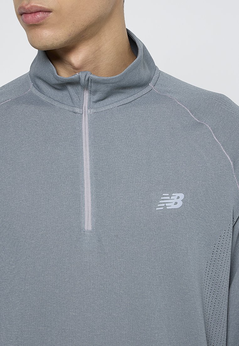 Hombre vestido con una camiseta deportiva de manga larga gris con cremallera hasta el pecho y el logo de New Balance en el pecho, además de un panel de tejido perforado en el costado.