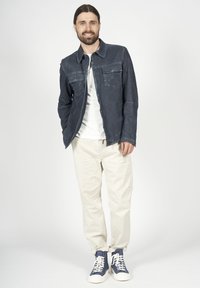 Mörkblå denimjacka med två framfickor, bärs över en vit skjorta. Krämfärgade byxor och marinblå och vita sneakers fulländar outfiten.