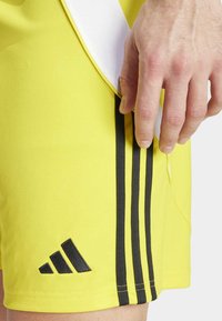 Geel sportshorts met een zwart drie-strepen ontwerp en een logo patch. De stof lijkt glad met een aansluitende snit en details van zijzakken.