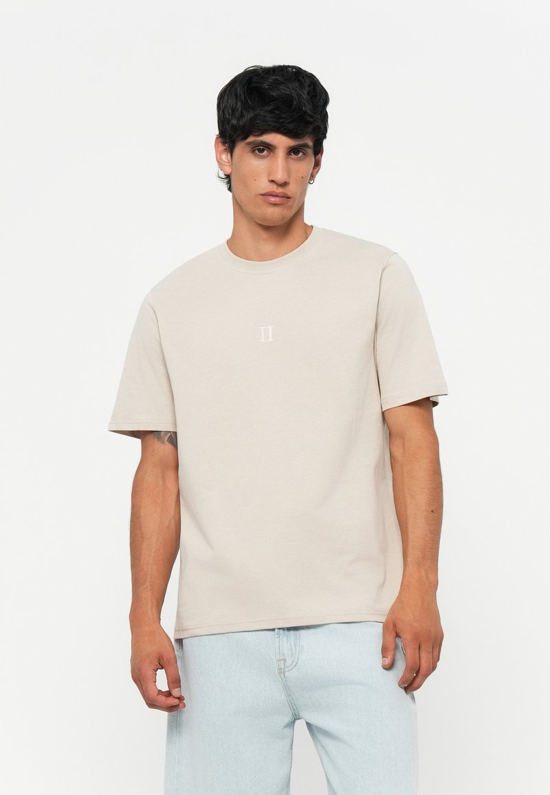 Beige cotton t-shirt with short sleeves, crew neck, and subtle "II" graphic in the center. Model wears light blue jeans.
