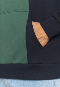 Main dans la poche d'un sweat à capuche bleu marine avec un panneau vert, bord-côte visible et jean en denim bleu clair en dessous.