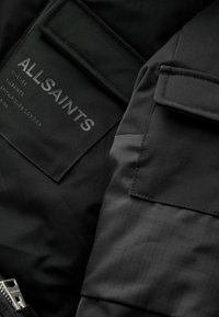 Chaqueta de tela negra con detalles mate y texturizados, cierre de cremallera y una etiqueta prominente con la marca "ALLSAINTS" en el bolsillo.
