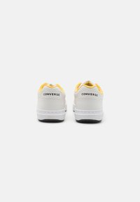 Bele tenisice z rumenimi detajli, gladek sintetični material, zaobljen nos, oblazinjen ovratnik in kontrastni črn gumijast podplat. Na peti je logotip "CONVERSE".