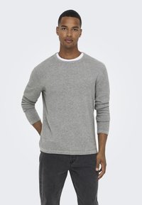 Maglietta grigia a maniche lunghe con un tessuto strutturato e un colletto rotondo bianco. Indossata con jeans scuri, la maglietta ha una vestibilità comoda e un design semplice.
