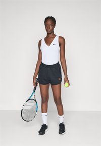 White ribbed tank top with a black Nike logo, black shorts with an accent logo, holding a tennis racket and ball, wearing black shoes.