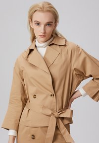 Manteau trench beige à double boutonnage avec ceinture à nouer, grand col et accents de boutons. Le tissu semble lisse avec une coupe décontractée.