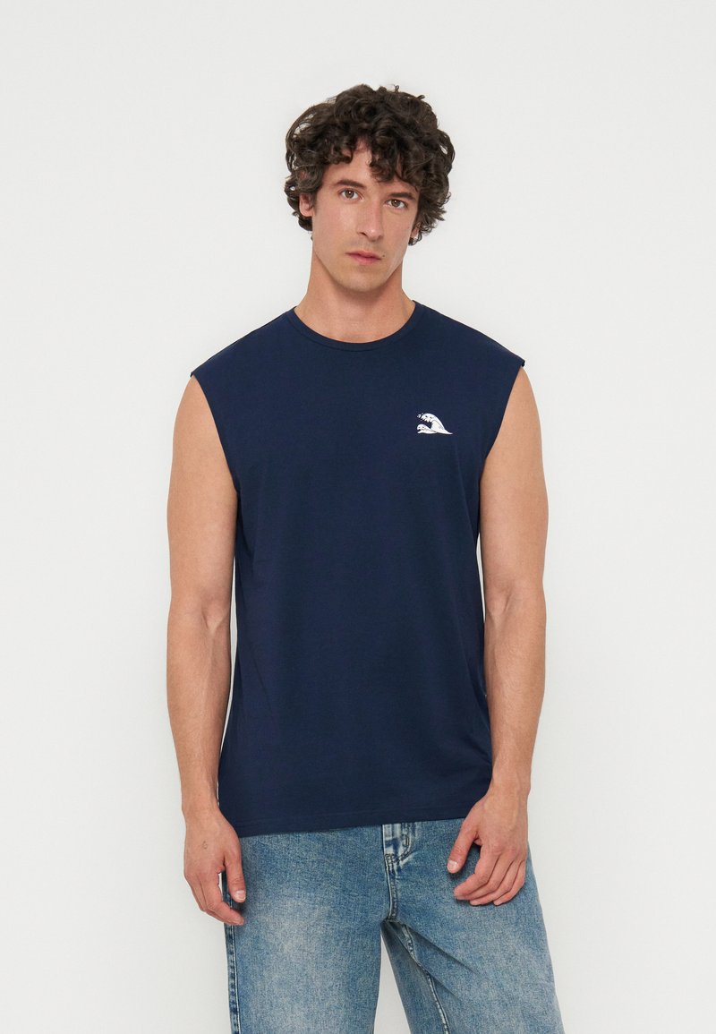 Homme aux cheveux bouclés portant un débardeur bleu marine avec un petit logo blanc en forme de vague sur la poitrine et un jean bleu clair, debout devant un fond blanc.