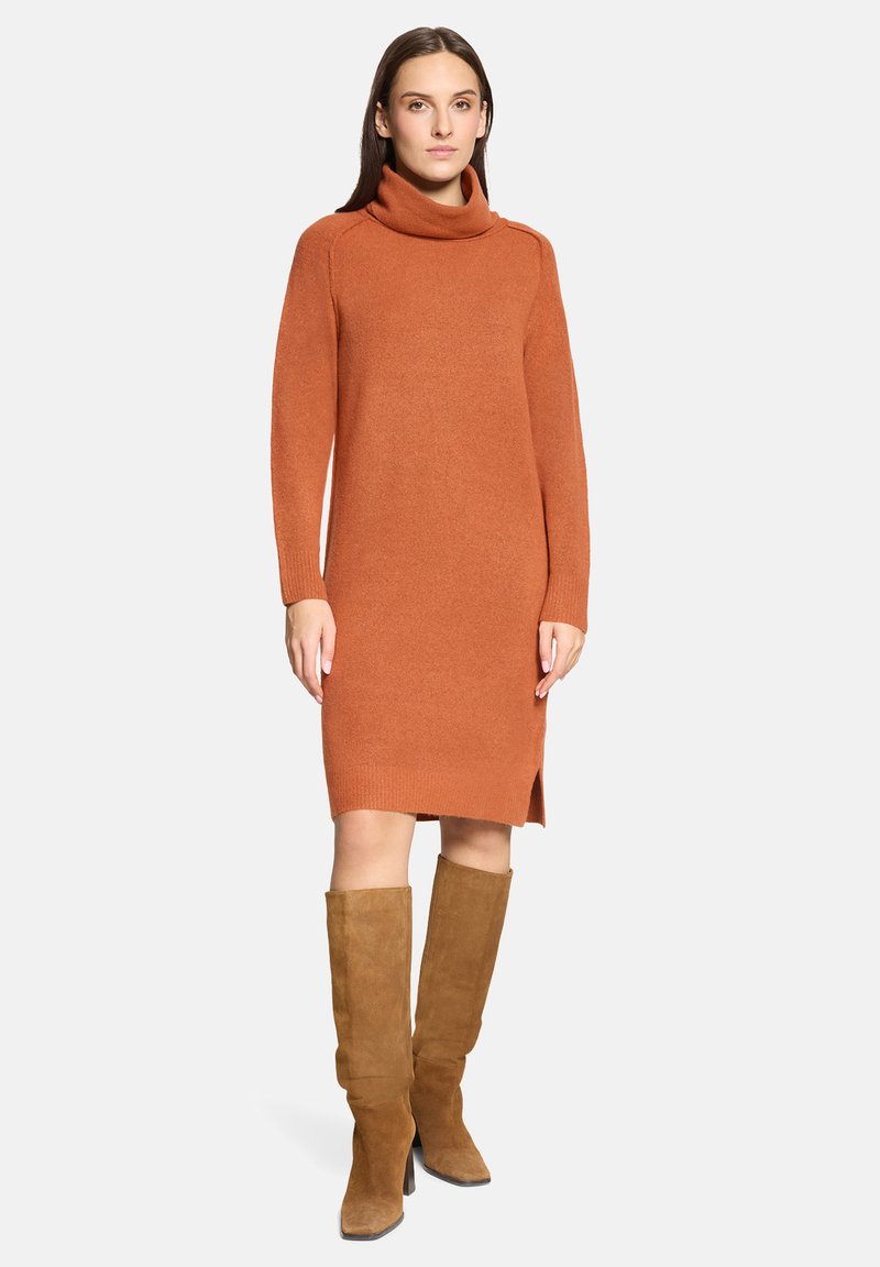 Femme debout portant une robe en maille col roulé orange brûlé à manches longues et des bottes hauteur genou en suède marron.