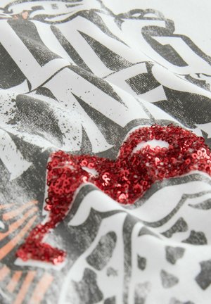 T-shirt en tissu blanc avec un texte graphique noir et un design en sequins rouges texturés. Texture douce avec un contraste de couleurs vif dans le motif.