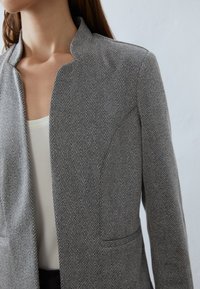 Blazer grigio a spina di pesce con colletto strutturato, dotato di due tasche frontali e un tessuto a finitura liscia. Sotto c'è un top bianco.