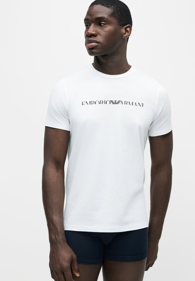 Άνδρας που φορά λευκό T-shirt Emporio Armani και σκούρα εσώρουχα, στέκεται και κοιτάζει στο πλάι μπροστά σε ένα απλό φωτεινό φόντο.