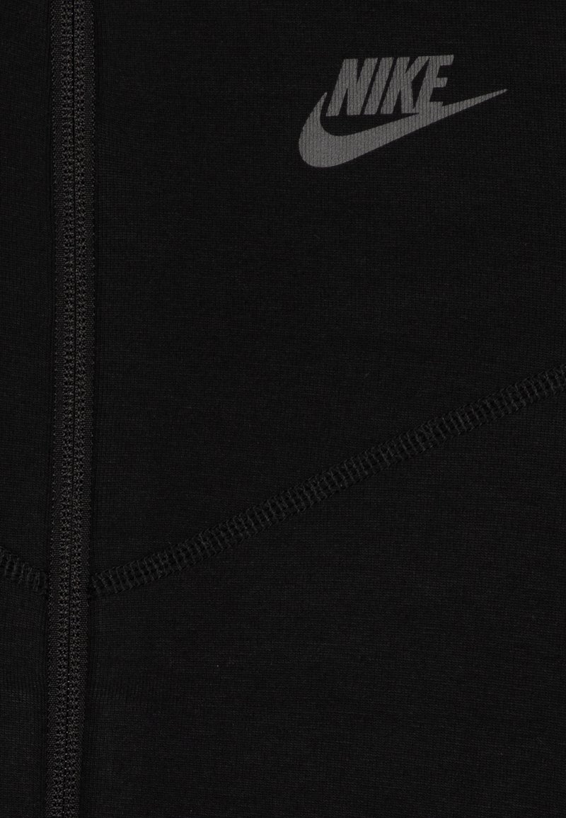 Hoodie noir à zip en tissu doux, arborant un logo Nike ton sur ton et des détails de couture contrastés. Texture lisse et design épuré.