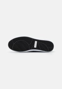 Semelle de sneaker en caoutchouc noir avec des motifs de grip texturés, présentant un accent blanc le long du bord et une petite étiquette de logo sur le côté.