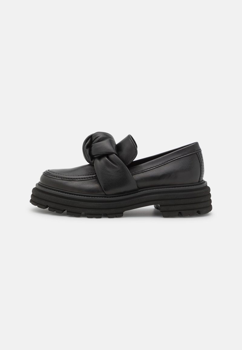 Mocassin en cuir noir avec semelle épaisse nervurée et grand nœud torsadé sur le dessus, présenté de profil sur fond blanc.