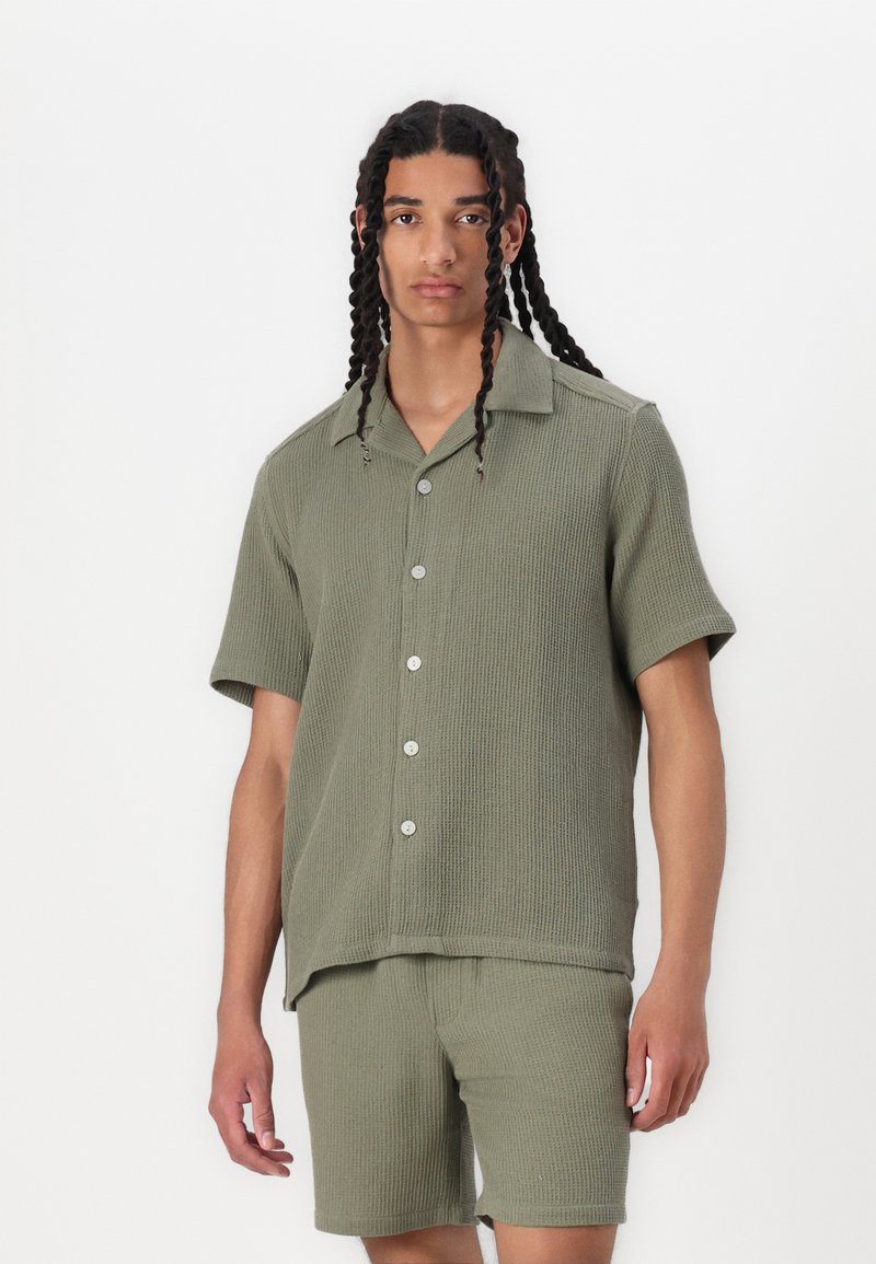 Olijfgroen, kortemouwens, geribbelde trui met een knoopsluiting aan de voorkant, gecombineerd met bijpassende shorts, voorzien van een relaxed fit en een iets oversized kraag.
