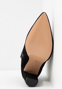 Stivaletto alla caviglia in suede nero con punta a punta rivolto verso l'alto, che mostra la suola liscia color nocciola con la taglia "4" vicino all'arco e una fibbia metallica sul lato.
