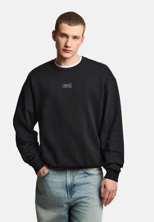 Młody mężczyzna o krótkich włosach, ubrany w czarną bluzę z małym logo z przodu, jasnoniebieskie dżinsy, stoi na jednolitym tle.