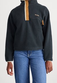 Pull en polaire gris foncé avec des accents beige, col montant et fermeture à boutons. Il comprend une poche poitrine zippée et un tissu texturé.