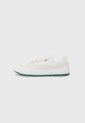 Zapatilla de ante beige con acentos blancos, forma redondeada y suela de goma verde. Presenta cordones y detalle de logo lateral.