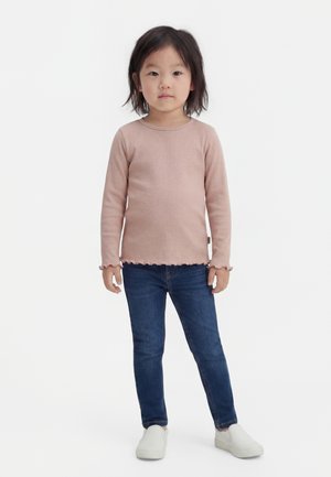 Otrok stoji, obrnjeni naprej, oblečen v roza dolg rokav, modre kavbojke in bele slip-on čevlje na belem ozadju.