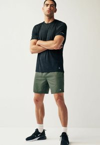 Fekete, rövidujjú sportos póló, finom csíkozott mintával; olíva zöld rövidnadrággal párosítva; a modell magabiztosan áll, keresztbe tett karokkal.