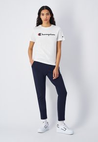 Biała bawełniana koszulka z logo "Champion" w czerni i czerwieni. Granatowe dopasowane spodnie z kieszeniami. Połącz z białymi sneakersami.