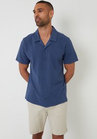 Korte mouwen overhemd met knopen in marineblauw, gemaakt van gladde stof, met een camp collar en vijf knopen aan de voorkant. Draagt met beige shorts.