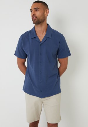 Camisa de botón de manga corta en azul marino, hecha de una tela suave, con un cuello tipo camp y cinco botones al frente. Llevado con pantalones cortos beige.